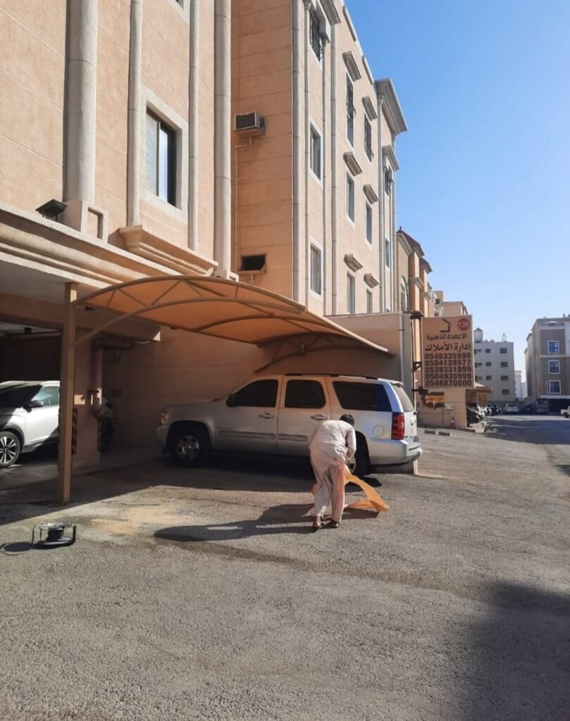 مظلة مواقف بتغطية فاتحة بجوار عمارة سكنية وسيارات أسفلها وعامل يعمل